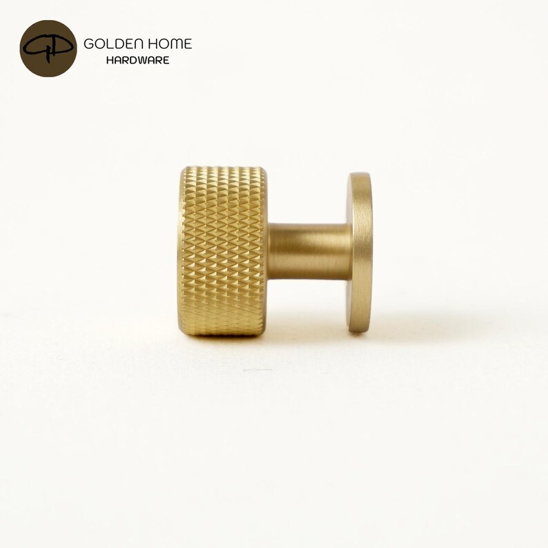 May include: A cylindrical brass cabinet knob with a textured, diamond-patterned knurled grip and a smooth, flat circular base. The hardware features a brushed gold finish and is presented alongside the text GOLDEN HOME HARDWARE.