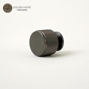 May include: A dark gray, cylindrical cabinet knob with a flat circular face and a textured, ribbed side surface. The knob features a metallic finish and is positioned horizontally. The logo for Golden Home Hardware is visible in the top left corner.