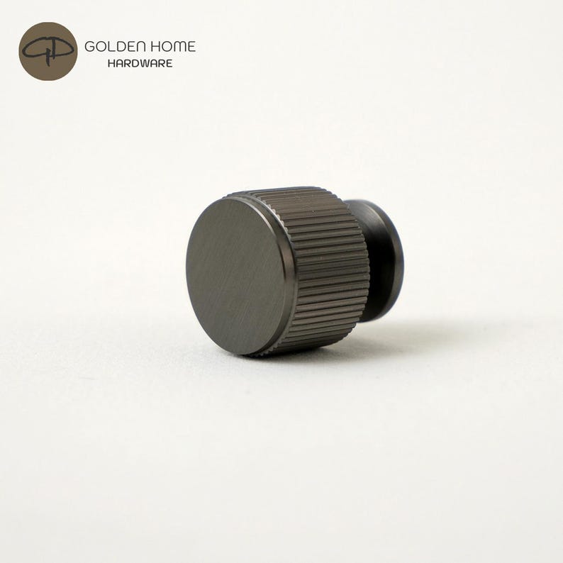 May include: A dark gray, cylindrical cabinet knob with a flat circular face and a textured, ribbed side surface. The knob features a metallic finish and is positioned horizontally. The logo for Golden Home Hardware is visible in the top left corner.