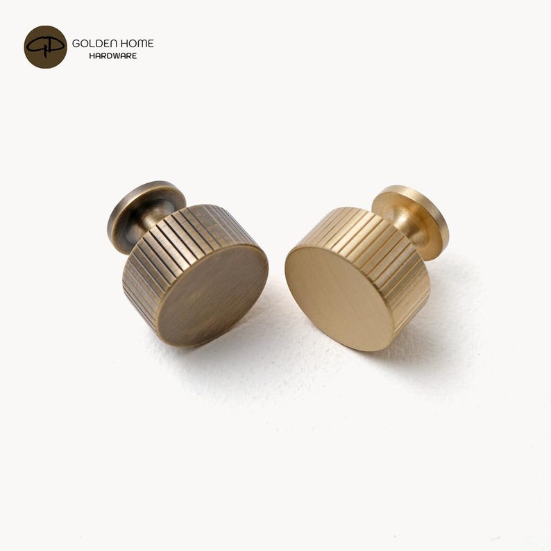 Op de afbeelding: Twee cilindrische kastknoppen met een verticale geribbelde textuur aan de zijkanten. E&eacute;n knop heeft een donkere antiek messing afwerking, terwijl de andere een heldere geborstelde gouden afwerking heeft. Beide knoppen hebben een ronde voet en een korte steel.