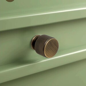 May include: A cylindrical cabinet knob with a brushed antique brass finish and vertical ribbed detailing, mounted on a sage green drawer front. The logo for Golden Home Hardware is visible in the upper left corner of the image.