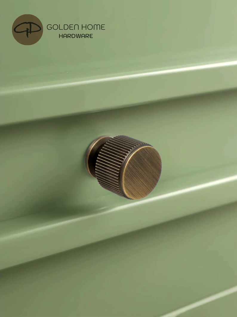 May include: A cylindrical cabinet knob with a brushed antique brass finish and vertical ribbed detailing, mounted on a sage green drawer front. The logo for Golden Home Hardware is visible in the upper left corner of the image.