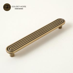 Puede incluir: Un tirador de mueble largo y rectangular en acabado de latón cepillado con extremos redondeados. La superficie presenta una serie de líneas negras paralelas y hundidas que siguen el contorno del tirador. El logo GOLDEN HOME HARDWARE es visible.