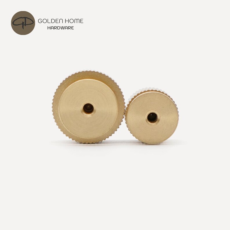 May include: Two circular brass knobs with a textured, knurled edge and a smooth, flat face featuring a centered hole. The hardware is branded with the text GOLDEN HOME HARDWARE in the upper left corner. These metallic components have a matte finish.