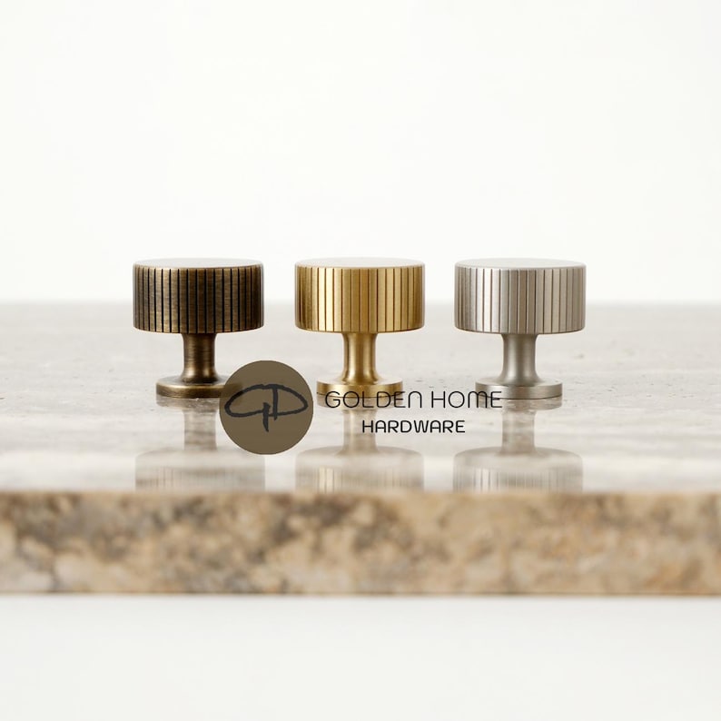 May include: Three cylindrical cabinet knobs with vertical ribbed detailing, arranged in a row. The left knob is dark bronze, the center is polished gold, and the right is brushed silver. The text GOLDEN HOME HARDWARE is visible in the center.