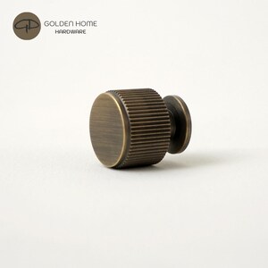 May include: A cylindrical cabinet knob with a brushed antique brass finish and a textured, ribbed edge. The knob features a flat circular face and a smaller circular base plate. The logo for Golden Home Hardware is visible in the top corner.