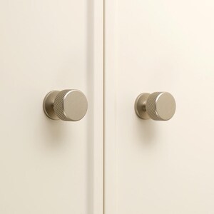 May include: Two circular cabinet knobs with a brushed champagne gold finish and a textured knurled edge. The knobs are mounted on a pair of off-white cabinet doors. The logo for Golden Home Hardware is visible in the top left corner of the image.