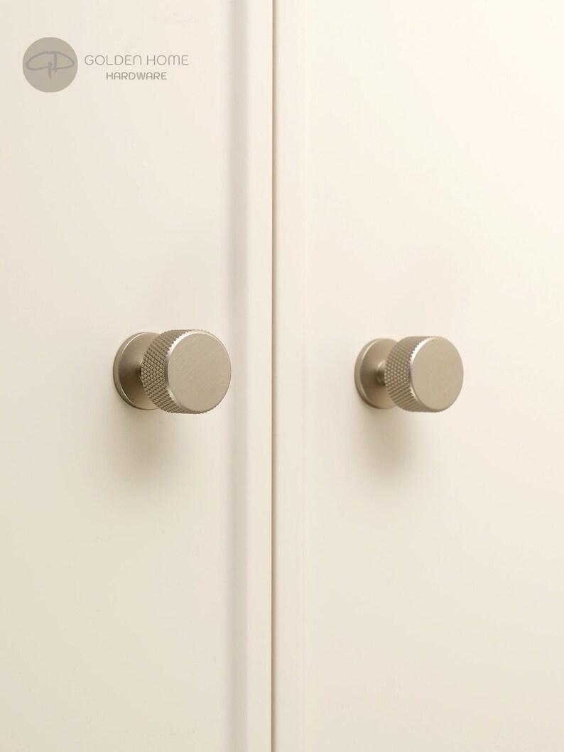 May include: Two circular cabinet knobs with a brushed champagne gold finish and a textured knurled edge. The knobs are mounted on a pair of off-white cabinet doors. The logo for Golden Home Hardware is visible in the top left corner of the image.