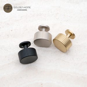 Op de afbeelding: Drie cilindrische kastknoppen van Golden Home Hardware, elk met een verticaal geribbelde textuur. De set bevat een matzwarte knop, een geborstelde zilveren knop en een geborstelde gouden knop, allemaal met een platte bovenkant en een kleine voet.
