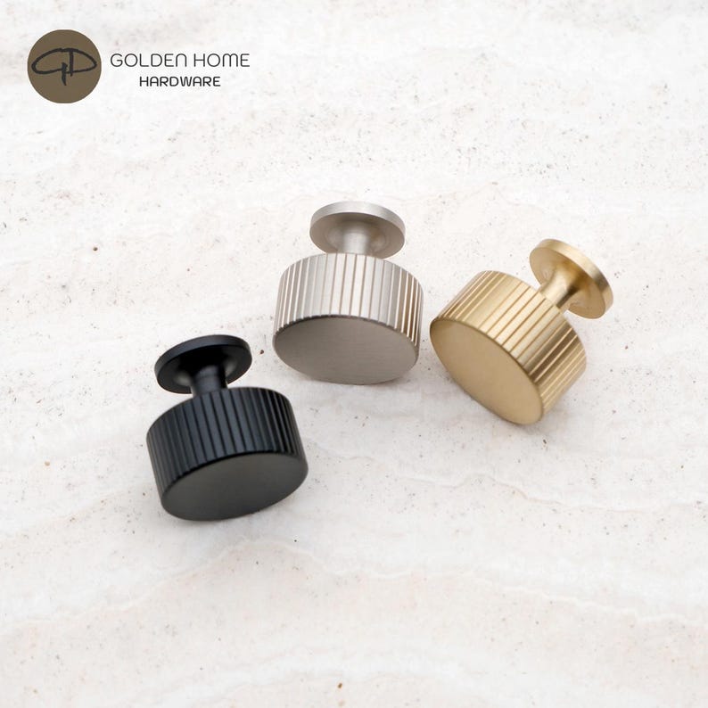 Op de afbeelding: Drie cilindrische kastknoppen van Golden Home Hardware, elk met een verticaal geribbelde textuur. De set bevat een matzwarte knop, een geborstelde zilveren knop en een geborstelde gouden knop, allemaal met een platte bovenkant en een kleine voet.