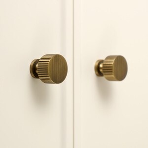 Op de afbeelding: Twee ronde kastknoppen met een geborstelde messing afwerking en een verticaal geribbelde textuur aan de zijkanten. De knoppen zijn gemonteerd op een effen gebroken wit oppervlak. Het logo Golden Home Hardware is zichtbaar linksboven.