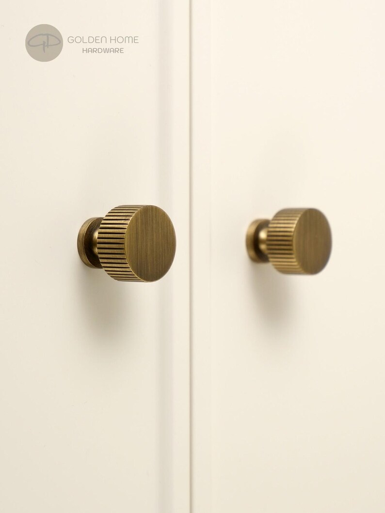 Op de afbeelding: Twee ronde kastknoppen met een geborstelde messing afwerking en een verticaal geribbelde textuur aan de zijkanten. De knoppen zijn gemonteerd op een effen gebroken wit oppervlak. Het logo Golden Home Hardware is zichtbaar linksboven.