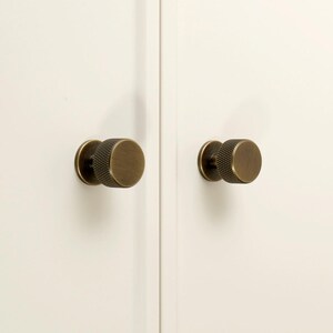 May include: Two circular cabinet knobs with a brushed antique brass finish and a textured knurled edge. The knobs are mounted on a plain white surface. The logo for Golden Home Hardware is visible in the top left corner of the image.