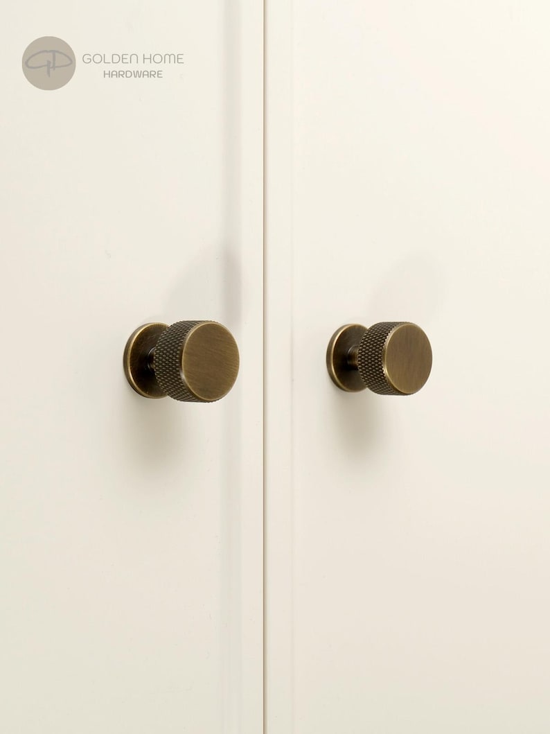 May include: Two circular cabinet knobs with a brushed antique brass finish and a textured knurled edge. The knobs are mounted on a plain white surface. The logo for Golden Home Hardware is visible in the top left corner of the image.