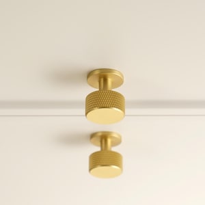 May include: Two cylindrical brass cabinet knobs with a textured knurled grip pattern on the sides and a smooth flat front face. The hardware is mounted on a plain white surface. The logo GOLDEN HOME HARDWARE is visible in the top left corner.