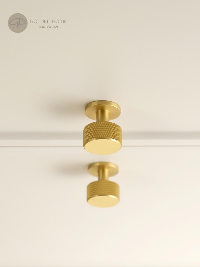 May include: Two cylindrical brass cabinet knobs with a textured knurled grip pattern on the sides and a smooth flat front face. The hardware is mounted on a plain white surface. The logo GOLDEN HOME HARDWARE is visible in the top left corner.