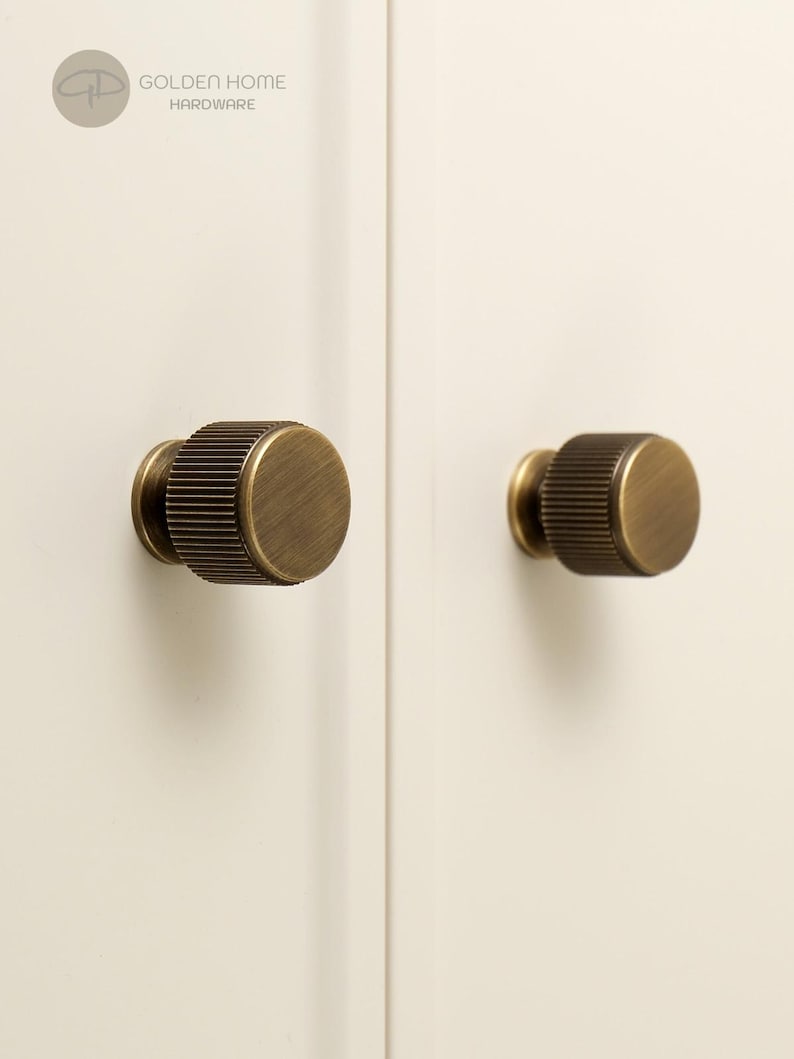 May include: Two cylindrical cabinet knobs with a brushed antique brass finish and a vertical ribbed texture on the sides. The knobs are mounted on a light cream-colored surface. A circular logo in the top left corner reads GOLDEN HOME HARDWARE.