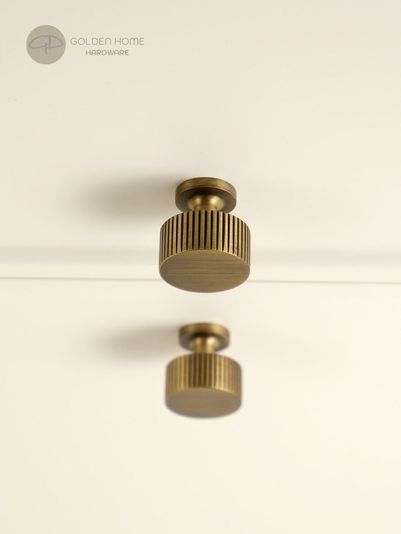 May include: Two cylindrical brass cabinet knobs with a vertical ribbed texture and a flat circular face. The knobs feature a brushed metallic finish and are mounted on a plain surface. The logo GOLDEN HOME HARDWARE is visible in the top left corner.