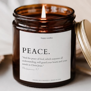 May include: A lit candle in an amber glass jar with a gold metal lid. The white label features the text "happy candles", "PEACE.", and the Bible verse "And the peace of God, which surpasses all understanding, will guard your hearts and your minds in Christ Jesus." Philippians 4:7.