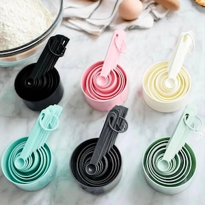 May include: Six sets of nested plastic measuring cups and spoons in black, pink, cream, teal, dark gray, and light green. Each set is held together by a circular ring, featuring graduated sizes for precise volume measurements in baking and cooking.