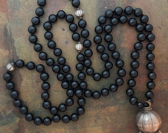 May include: A black onyx mala necklace with silver accents and a large silver pendant. The necklace is made of round beads and is approximately 108 beads long.