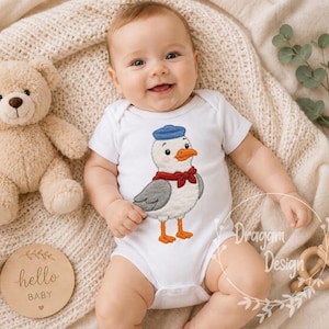 Könnte beinhalten: Ein weißer, kurzärmeliger Babybody mit einer Stickerei einer Möwe, die eine blaue Matrosenmütze und eine rote Fliege trägt. Die Möwe hat graue Flügel, einen orangefarbenen Schnabel und orangefarbene Füße.