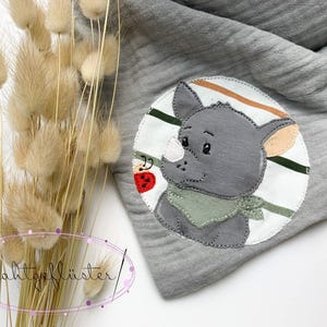 Puede incluir: Una bufanda de muselina gris con un parche circular que muestra un rinoceronte de dibujos animados con un pañuelo verde. El parche incluye una pequeña mariquita roja y rayas verdes horizontales. El texto "Wahlgeflüster" es visible en la esquina.