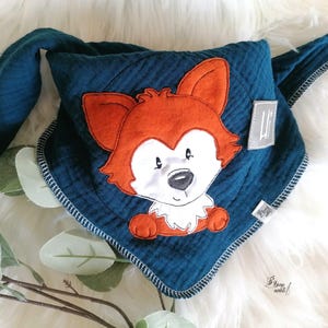 May include: A dark teal triangular bandana bib featuring a central embroidered orange and white fox character. The bib has a white stitched edge and a small grey fabric tag with the text "Hi" and another smaller tag with illegible text.