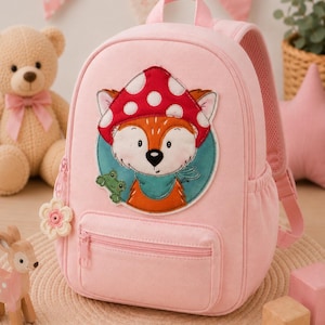 Könnte beinhalten: Ein hellrosa Rucksack mit einem zentralen gestickten Aufnäher eines Fuchses, der einen roten Pilzhut mit weißen Punkten und einen türkisfarbenen Schal trägt, begleitet von einem kleinen grünen Frosch. Der Rucksack hat eine Vordertasche mit Reißverschluss und einen blumenförmigen Zipper.
