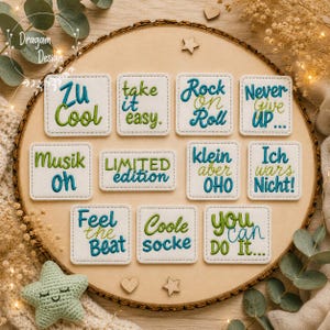 Peut inclure: Une collection de onze écussons en tissu blanc, carrés et rectangulaires, avec du texte brodé en fil bleu et vert. Les phrases incluent Zu Cool, take it easy, Rock on Roll, Never Give UP, Musik oh, LIMITED edition, klein aber OHO, Ich wars Nicht, Feel the Beat, Coole socke et YOU can DO It.