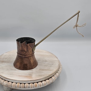 May include: A copper coffee pot with a wide base, a narrow neck, and a flared rim. It features a long, thin, angled metal handle with a piece of brown twine tied to the end. The exterior surface of the pot shows a weathered, dark patina.