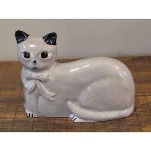 May include: A ceramic cat figurine in a reclining position with a light gray speckled finish. The cat features black painted ears, large almond-shaped eyes, a small nose, and a sculpted bow tied around its neck. The surface has a glossy glaze.