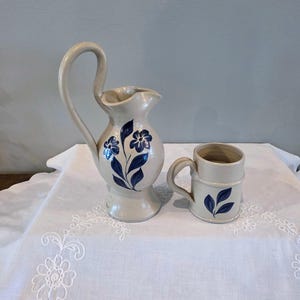 May include: A beige ceramic pitcher with a tall, curved handle and a matching mug, both decorated with hand-painted dark blue floral and leaf motifs. The pitcher features a flared base and a spout, while the mug has a simple cylindrical shape.