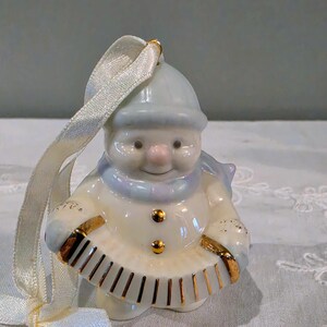 May include: A glossy porcelain snowman ornament wearing a light blue hat and scarf, featuring two gold buttons and holding a white accordion with gold trim. A cream-coloured satin ribbon is attached to the top for hanging this festive decoration.
