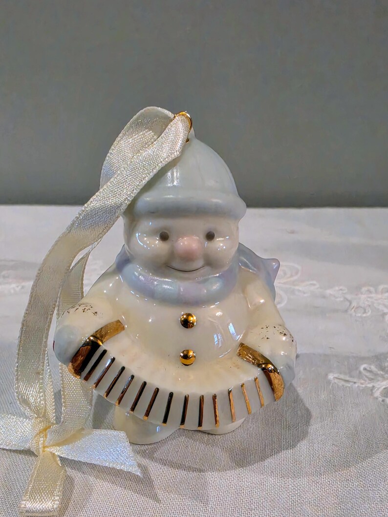May include: A glossy porcelain snowman ornament wearing a light blue hat and scarf, featuring two gold buttons and holding a white accordion with gold trim. A cream-coloured satin ribbon is attached to the top for hanging this festive decoration.