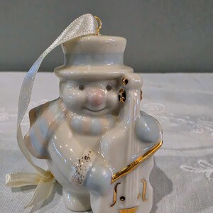 May include: A glossy ceramic snowman ornament wearing a top hat and a striped scarf, playing a double bass. The snowman is white with soft blue and pink accents, featuring gold-painted details on the instrument and a cream-coloured ribbon loop.