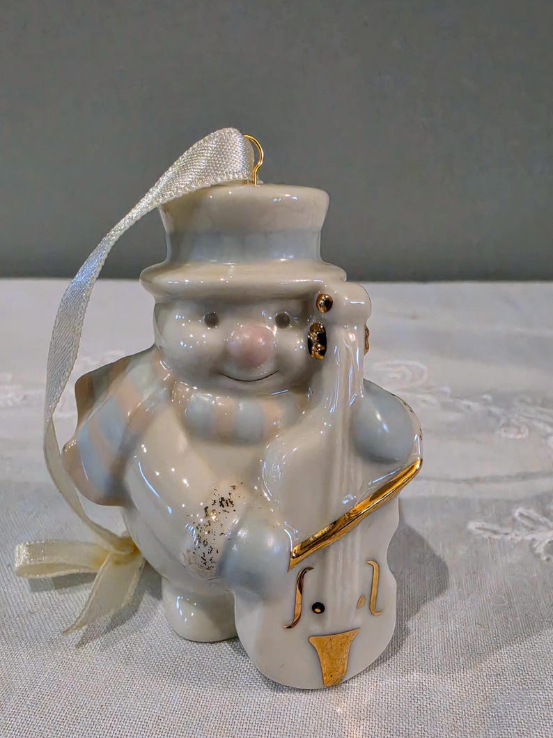 May include: A glossy ceramic snowman ornament wearing a top hat and a striped scarf, playing a double bass. The snowman is white with soft blue and pink accents, featuring gold-painted details on the instrument and a cream-coloured ribbon loop.