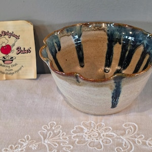 May include: A ceramic baking dish with a scalloped rim, featuring a tan base and dark blue glaze drips running down the interior and exterior. Beside it is a paper label reading The Original Apple Baker Baking Dish from Christian Ridge Pottery.