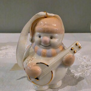 May include: A ceramic snowman ornament featuring a peach-coloured hat and scarf, playing a banjo with gold-tone accents. The figure has a glossy white finish and includes a cream-coloured fabric ribbon loop attached to the top for hanging purposes.