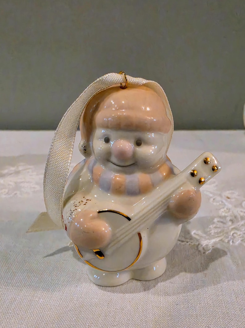 May include: A ceramic snowman ornament featuring a peach-coloured hat and scarf, playing a banjo with gold-tone accents. The figure has a glossy white finish and includes a cream-coloured fabric ribbon loop attached to the top for hanging purposes.