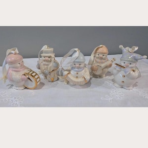 May include: A set of five porcelain snowman ornaments, each featuring gold accents and a white ribbon for hanging. The figures are depicted playing various musical instruments, including a drum, a banjo, an accordion, and a flute, with pastel-coloured hats.