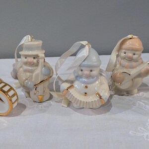 May include: A set of five porcelain snowman ornaments, each featuring gold accents and a white ribbon for hanging. The figures include snowmen playing a drum, a cello, an accordion, a banjo, and a flute, all finished in soft pastel colours.