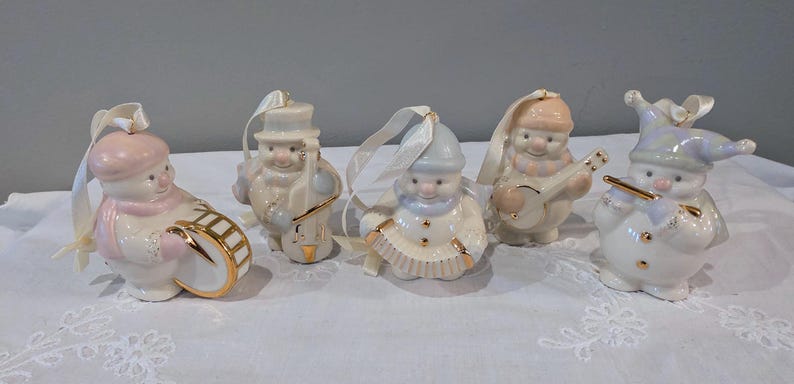 May include: A set of five porcelain snowman ornaments, each featuring gold accents and a white ribbon for hanging. The figures include snowmen playing a drum, a cello, an accordion, a banjo, and a flute, all finished in soft pastel colours.