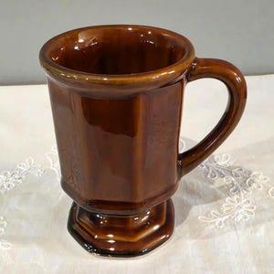 May include: A glossy, dark brown ceramic mug featuring a faceted, octagonal body and a matching octagonal pedestal base. The mug has a smooth, reflective finish and a curved handle extending from the side, showcasing a solid, uniform color.