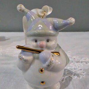 May include: A ceramic snowman ornament wearing a pastel purple and green jester hat with gold glitter accents. The snowman holds a shiny gold flute, has a pink nose, and features two small gold buttons on its round white body with a white ribbon loop.