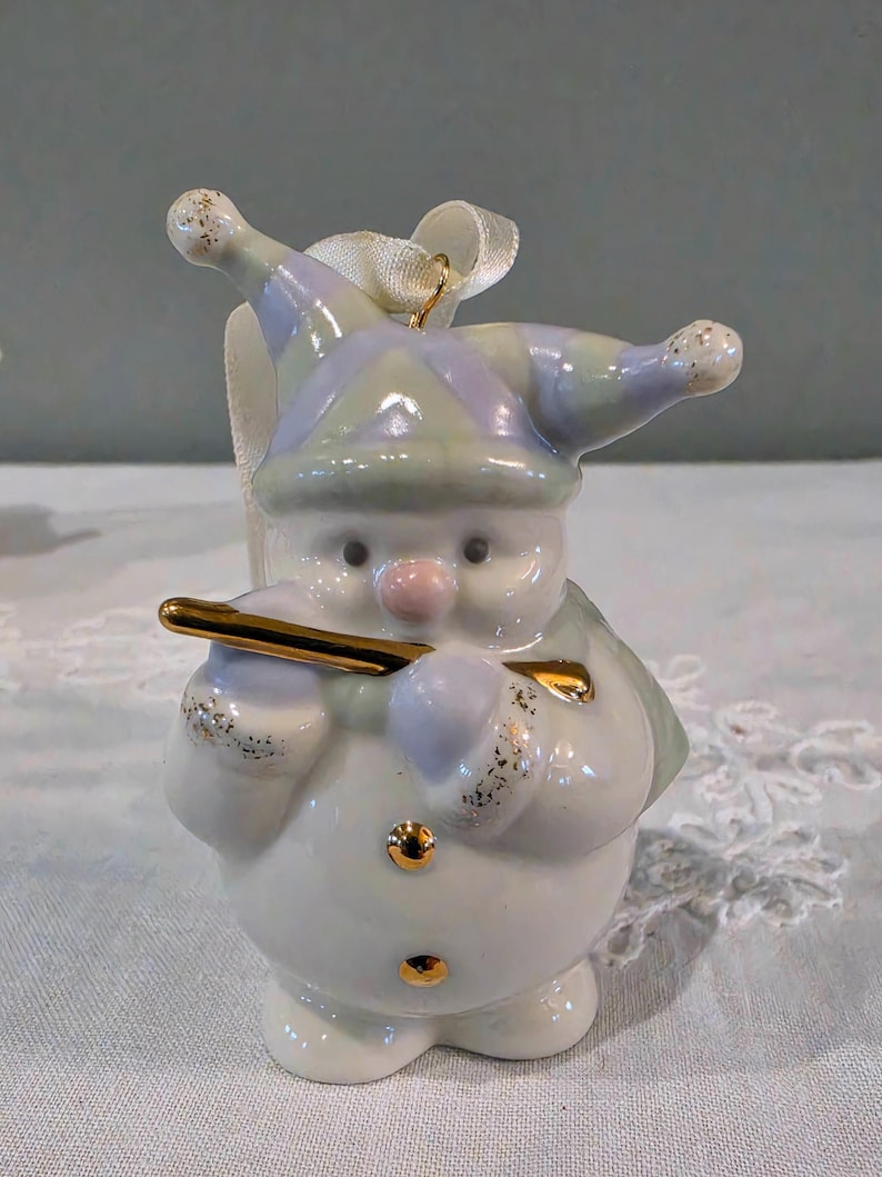 May include: A ceramic snowman ornament wearing a pastel purple and green jester hat with gold glitter accents. The snowman holds a shiny gold flute, has a pink nose, and features two small gold buttons on its round white body with a white ribbon loop.