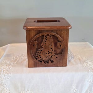 May include: A square wooden tissue box cover featuring a dark brown finish and a circular cutout on the front panel. The cutout displays an intricate, carved butterfly design with detailed wing patterns. The top has a rectangular opening.