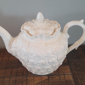 May include: A cream-colored ceramic teapot featuring a textured pineapple-inspired surface pattern. The teapot includes a matching lid with a small finial, a curved spout, and a handle. The overall design is monochromatic with a glossy finish.