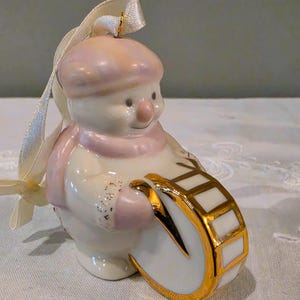 May include: A ceramic snowman ornament featuring a soft pink hat and scarf, holding a white drum with gold trim. The snowman has a glossy finish with subtle gold glitter accents on the mittens and includes a cream-coloured ribbon for hanging.