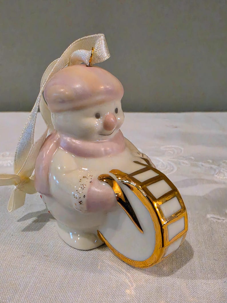 May include: A ceramic snowman ornament featuring a soft pink hat and scarf, holding a white drum with gold trim. The snowman has a glossy finish with subtle gold glitter accents on the mittens and includes a cream-coloured ribbon for hanging.